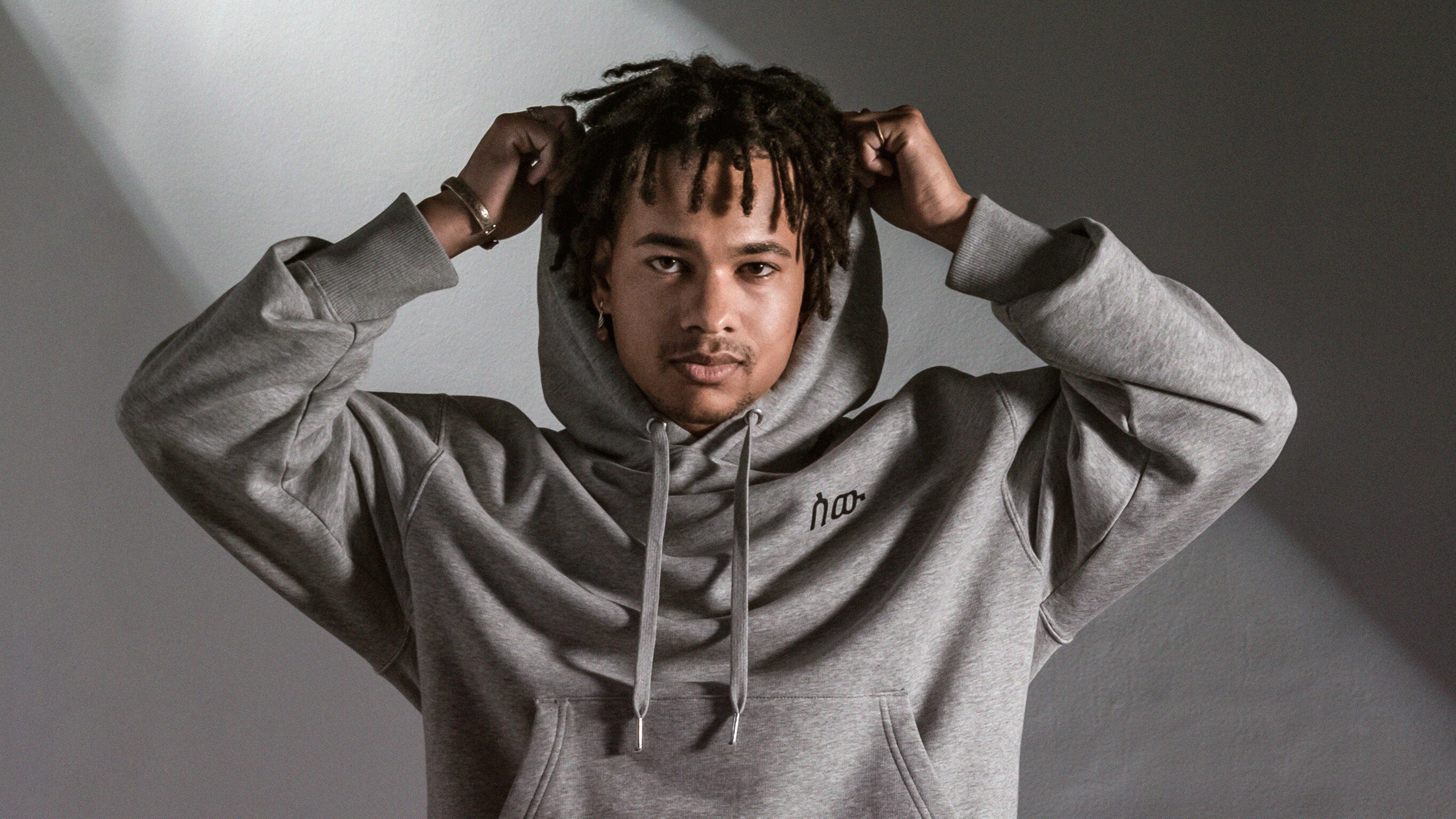 Ein junger Mann mit dunklen Locken und Bart steht frontal vor einer hellgrauen Wand. Er trägt einen grauen Hoodie mit dem schwarzen „∞oo“-Logo auf der Brust und zieht sich mit beiden Händen gerade die Kapuze über den Kopf. Das Licht fällt seitlich auf ihn und erzeugt einen weichen Schatteneffekt, der die Szene ruhig und fokussiert wirken lässt.