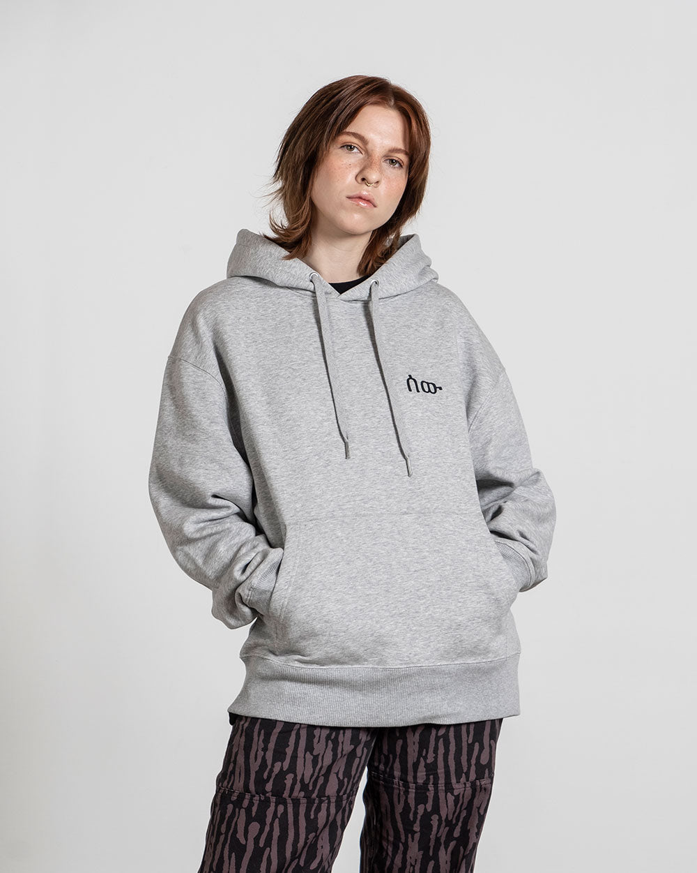 Grauer Kapuzenpullover mit lockerer Passform, Kängurutasche und schwarzem Frontprint eines minimalistischen Symbols auf der Brust, getragen von einer Person mit rötlich-braunen Haaren und dunkler gemusterter Hose.