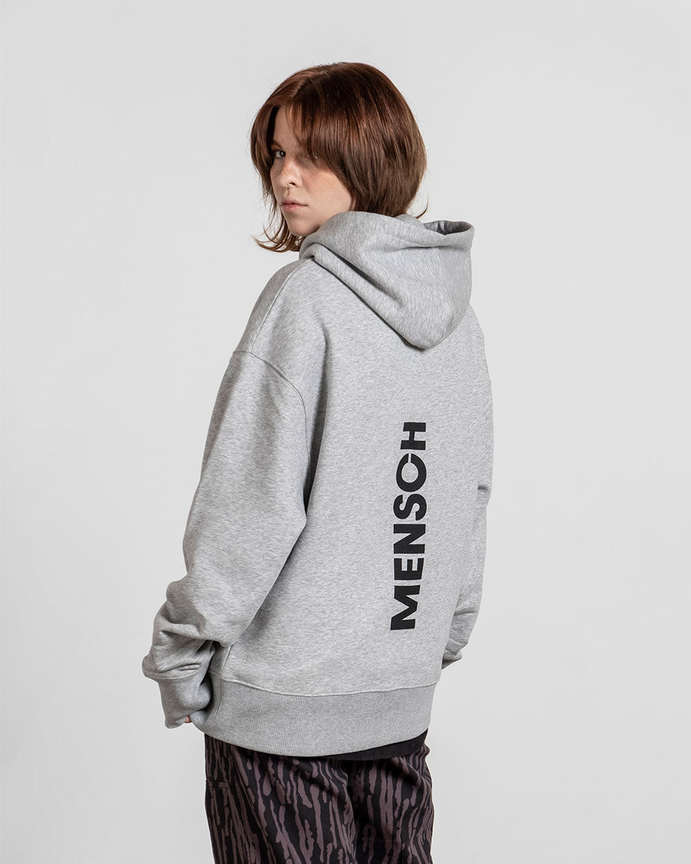 Rückseite des grauen Hoodies mit vertikalem schwarzen Aufdruck „MENSCH“ entlang der Wirbelsäule; die Kapuze ist hochgezogen, das Model blickt über die Schulter.