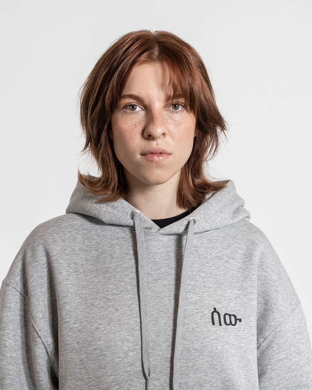 Grauer Hoodie mit lockerem Schnitt und mittig platziertem schwarzen Brustmotiv aus abstrakten Zeichen, getragen von einer Person mit rötlich-braunen Haaren.