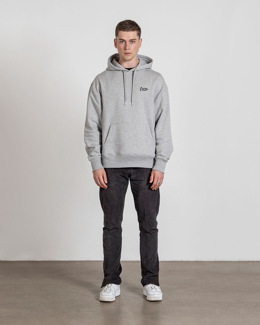 Grauer Hoodie mit Frontprint in Kombination mit dunkler Jeans und weißen Sneakern; das Model steht frontal zur Kamera mit neutralem Gesichtsausdruck.