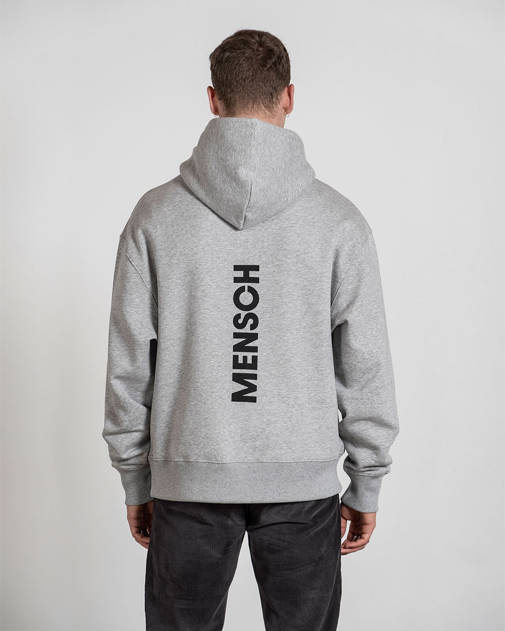 Grauer Hoodie mit großem schwarzem „MENSCH“-Print auf der Rückseite, getragen von einer Person mit kurz geschnittenem Haar; die Kapuze liegt auf dem Rücken.