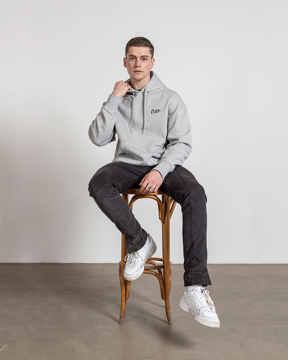 Grauer Kapuzenpullover mit dunkler Hose und weißen Sneakern, getragen von einem Model, das auf einem Holzstuhl sitzt und mit einer Hand den Kragen hält.