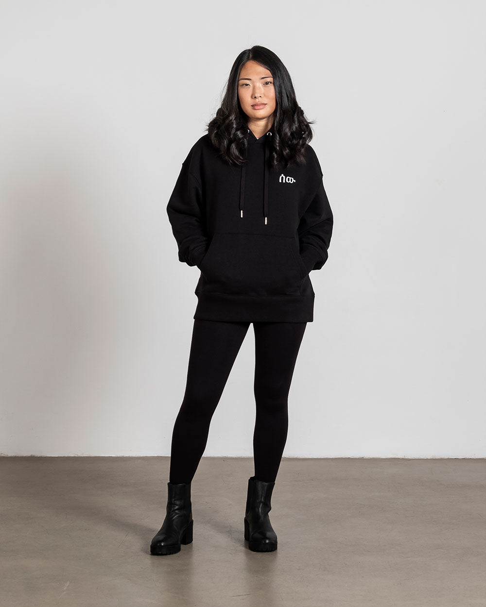 Vorderansicht eines schwarzen Hoodies mit weißer, minimalistisch gestickter Symbolschrift auf der linken Brust. Getragen von einer stehenden Person mit den Händen in der Kängurutasche.