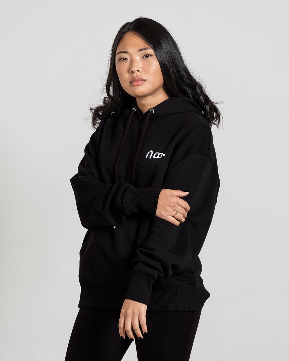 Schwarz gekleidete Person trägt den Hoodie frontal und hält einen Arm locker vor dem Körper. Die weiße Stickerei des Logos ist deutlich sichtbar.