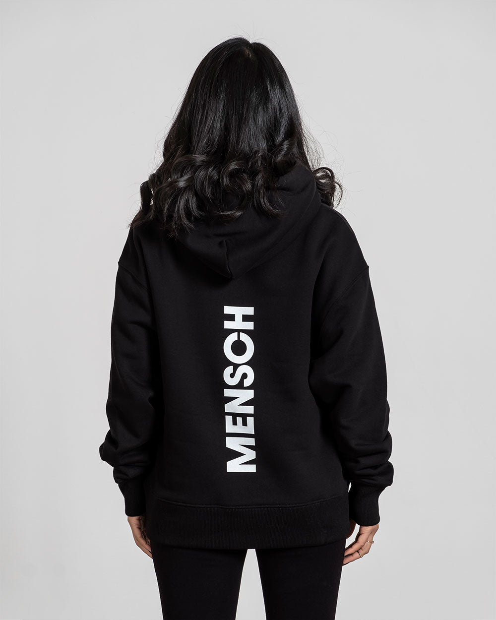 Rückansicht des schwarzen Hoodies mit vertikalem, großem weißen Schriftzug „MENSCH“ in der Mitte des Rückens.