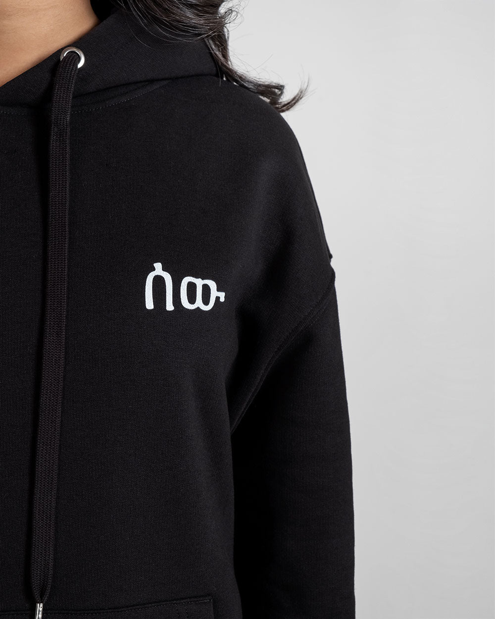 Detailaufnahme der weißen Frontstickerei des schwarzen Hoodies. Der Schriftzug ist stilisiert und modern.