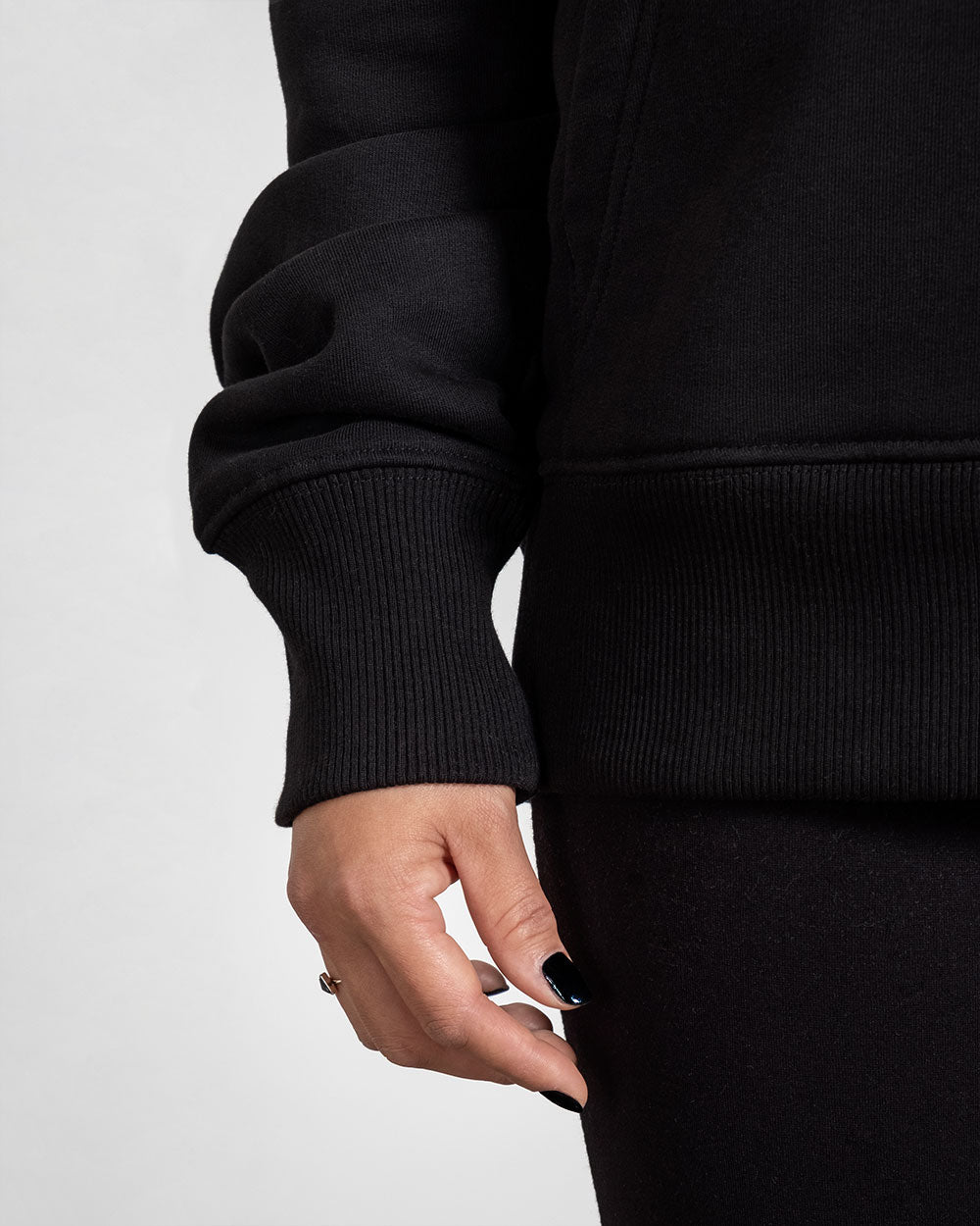 Detailaufnahme der rechten Ärmelpartie mit geripptem Bündchen des schwarzen Hoodies, leicht über der Hand fallend.