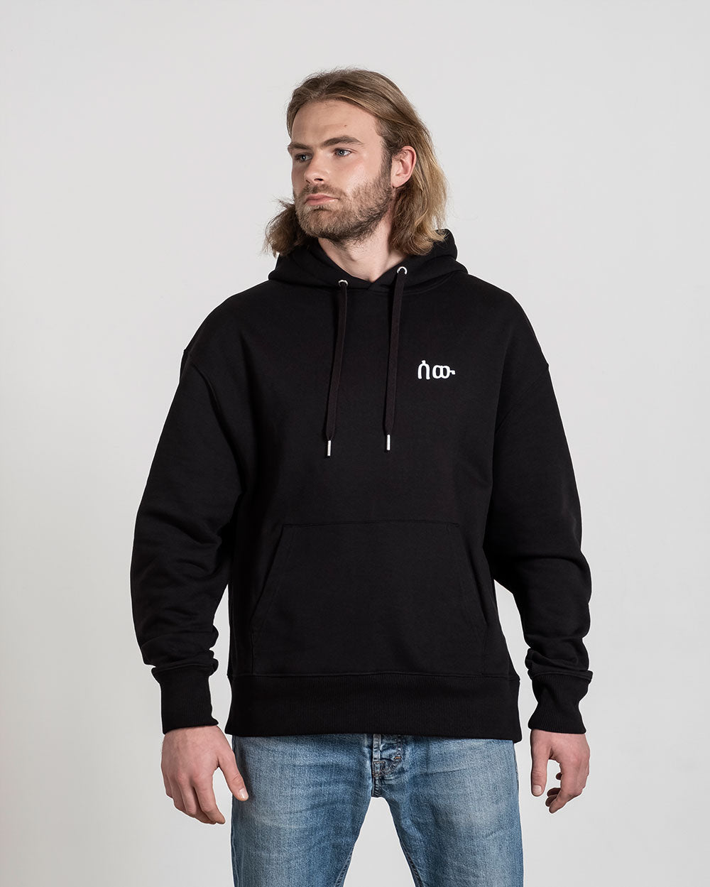 Frontalansicht eines schwarzen Hoodies mit weißer Bruststickerei, getragen von einer Person in Jeans. Die Kapuze ist locker nach hinten gelegt.