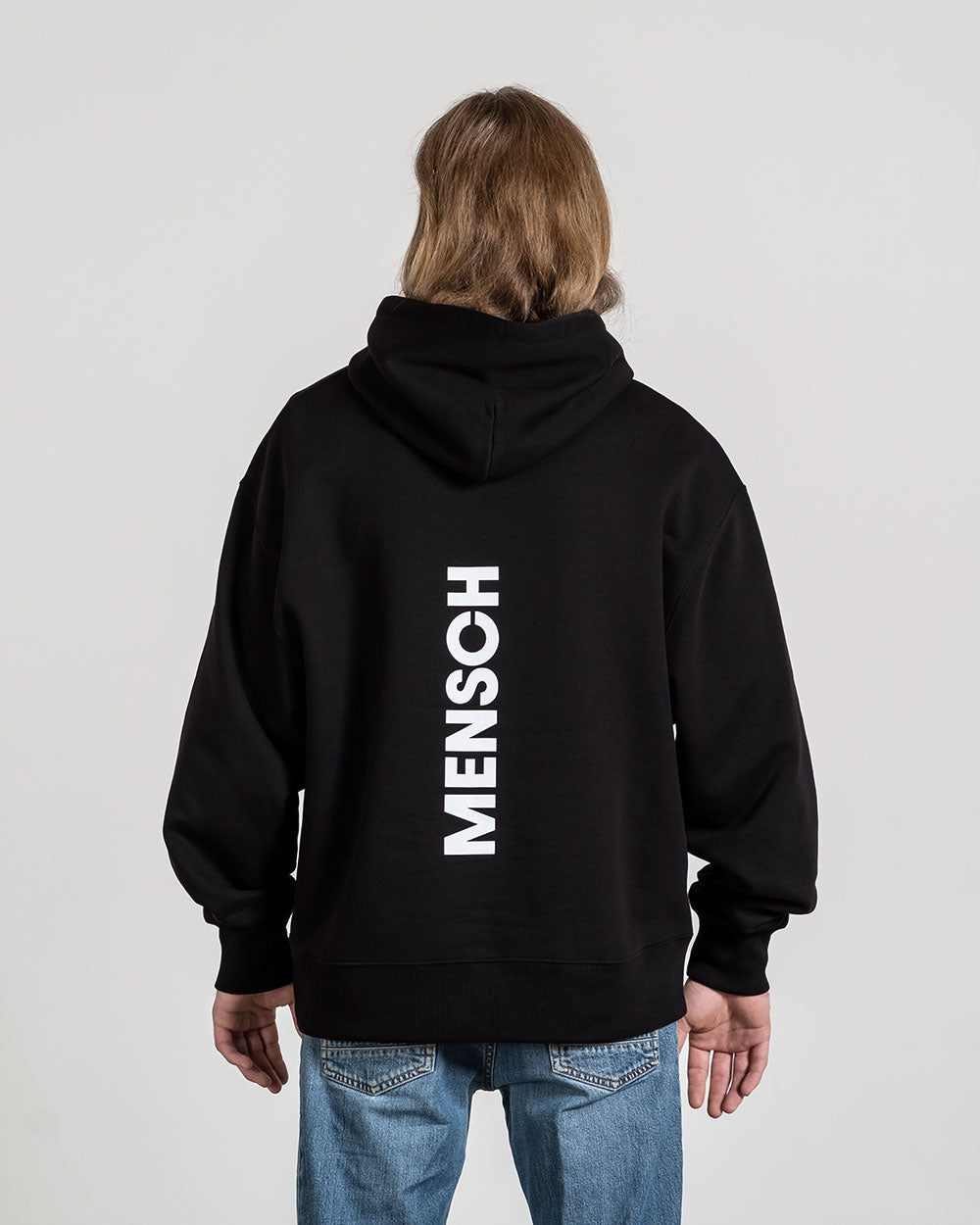 Rückansicht des schwarzen Hoodies mit vertikalem weißen „MENSCH“-Schriftzug, getragen von einer Person mit mittellangem Haar.