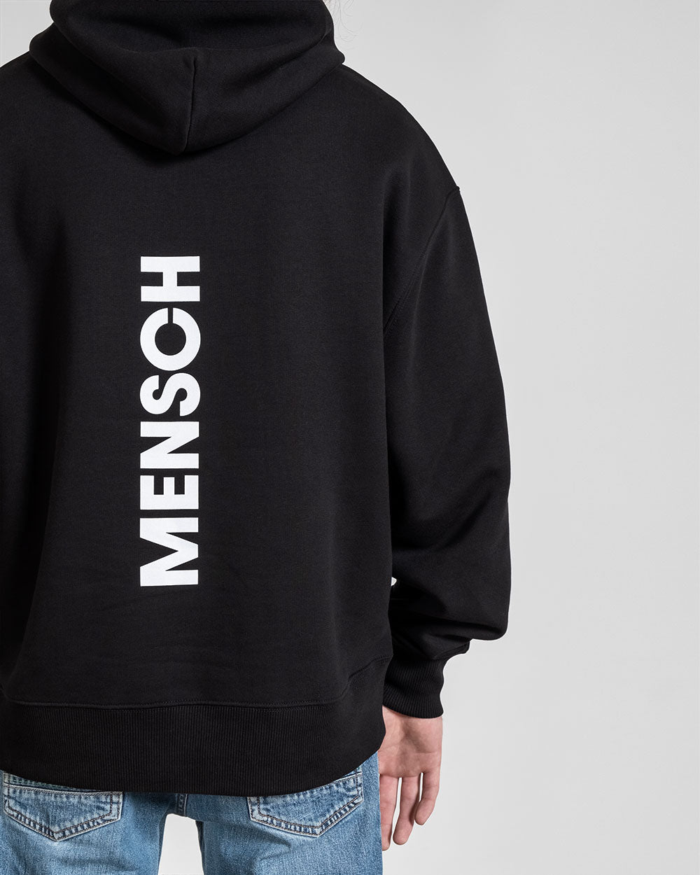 Nahaufnahme des Rückens: weißer „MENSCH“-Schriftzug auf schwarzem Stoff, zentriert und vertikal platziert, direkt unterhalb der Kapuze.