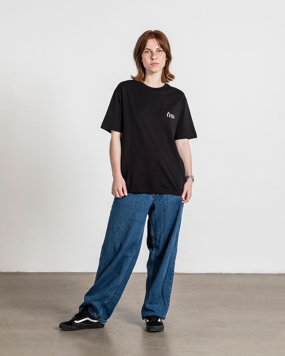Schwarzes T-Shirt mit weißem Brustlogo, locker an einer Person mit Jeans getragen, frontal aufgenommen.