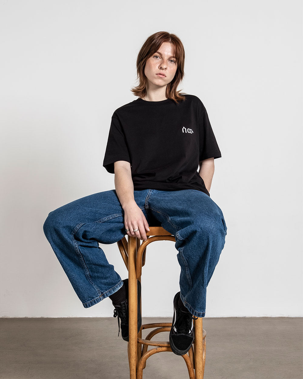 Schwarzes T-Shirt mit weißem Logo auf der Brust, locker an einer sitzenden Person getragen.