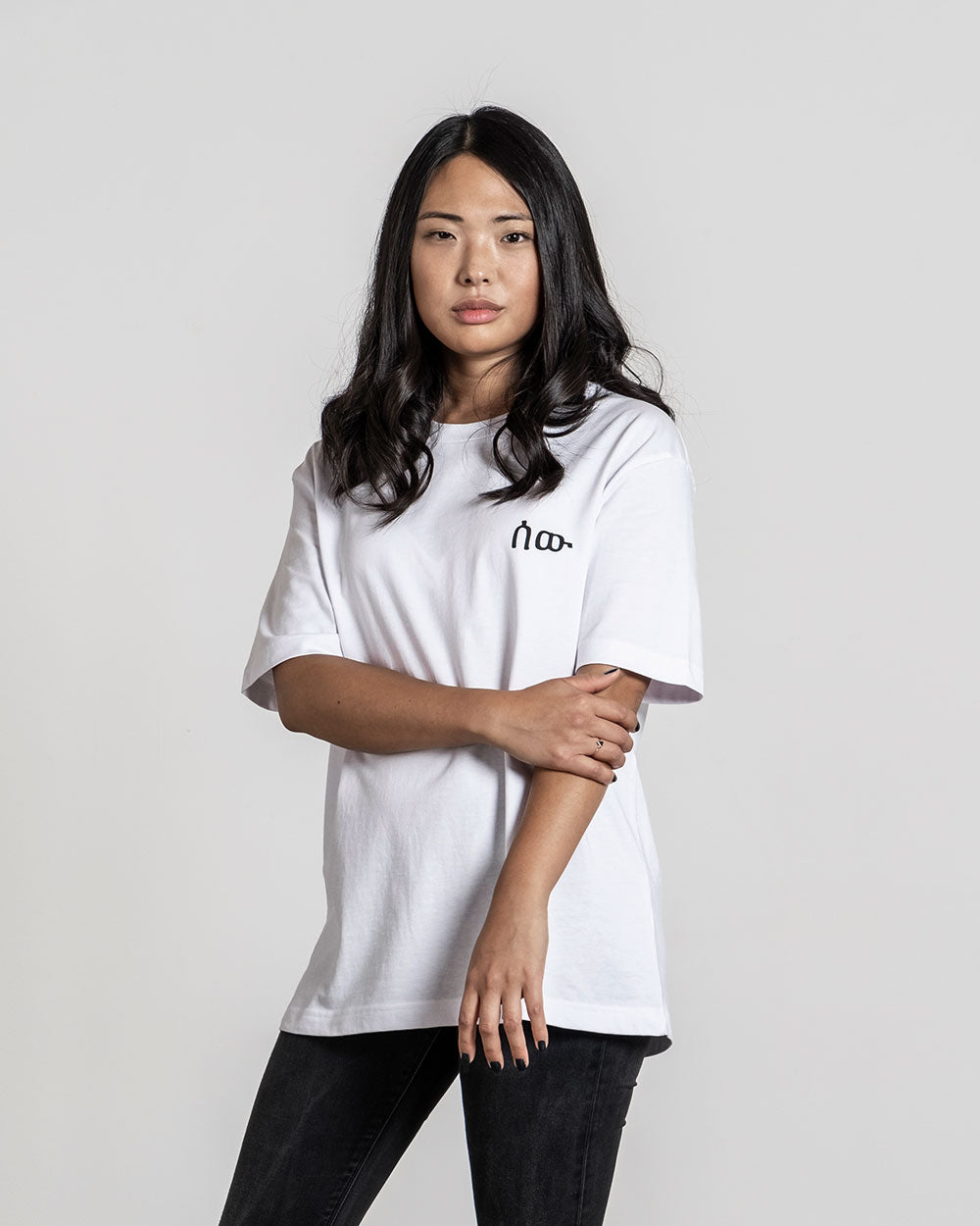 Weißes T-Shirt mit schwarzem Frontprint eines stilisierten Schriftzugs auf der linken Brust, getragen von einer Person mit dunklem Haar, Blick frontal.