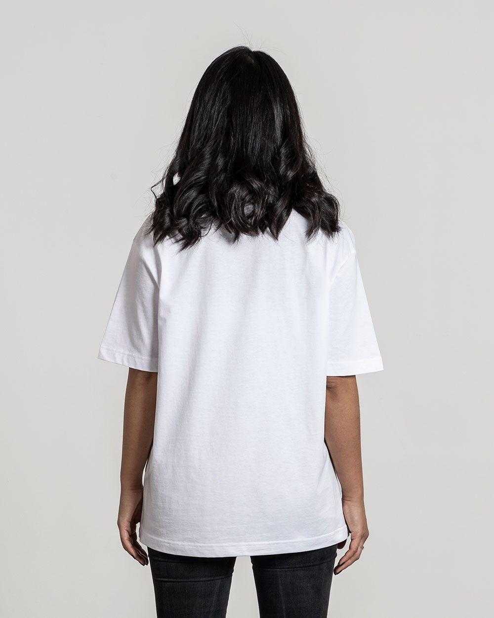 Rückansicht eines weißen T-Shirts ohne sichtbaren Aufdruck im Schulterbereich, getragen von einer Person mit offenen, dunklen Haaren.