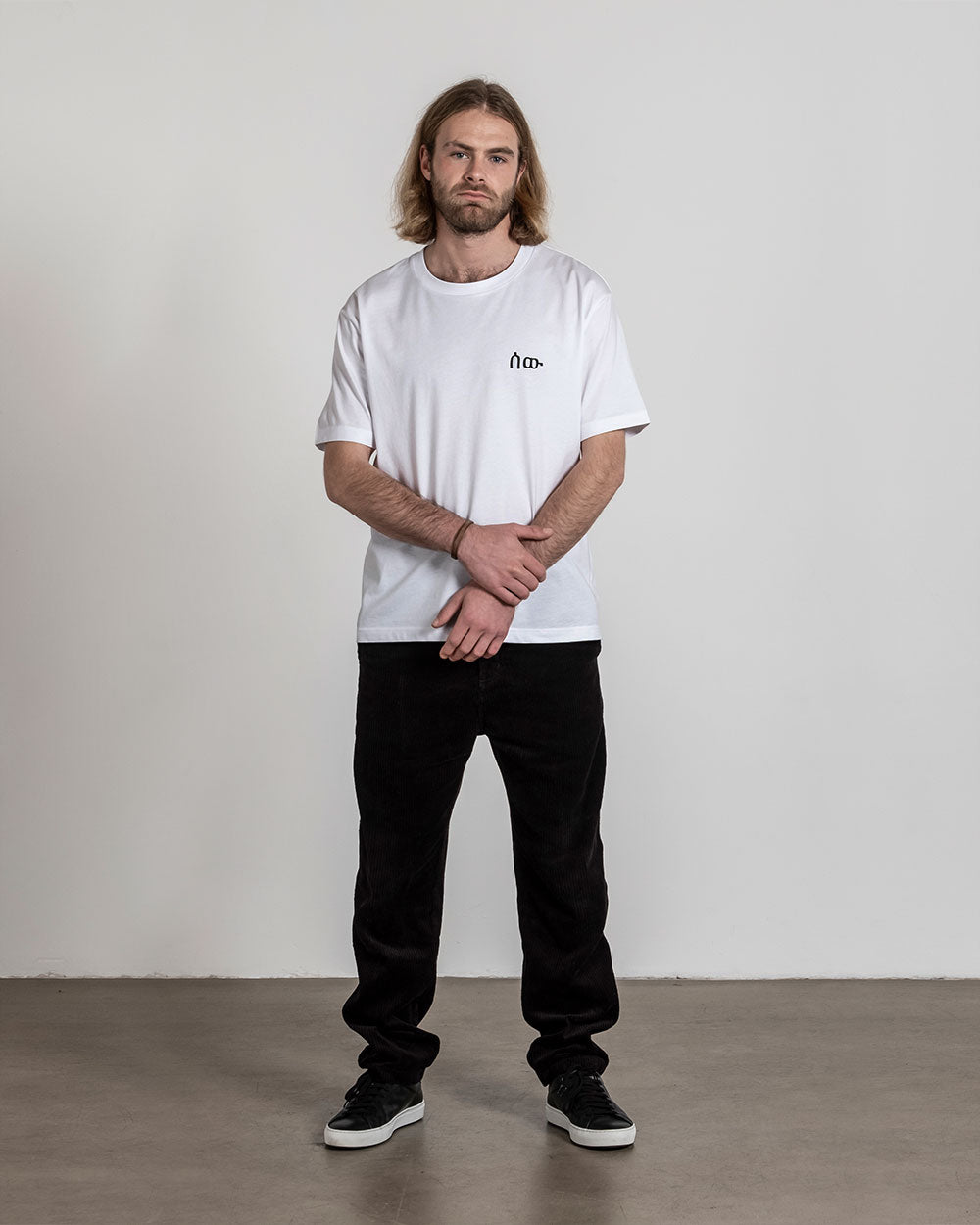 Weißes T-Shirt mit schwarzem Frontprint auf der linken Brust, getragen von einer Person mit langen Haaren und Bart, stehend mit verschränkten Armen.