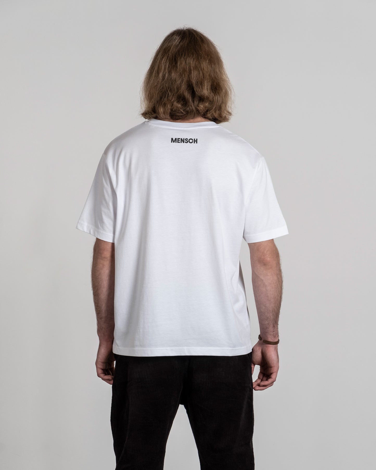 Rückansicht eines weißen T-Shirts mit kleinem, schwarzen Rückendruck „MENSCH“ im oberen Schulterbereich, getragen von einer Person mit mittellangem Haar.