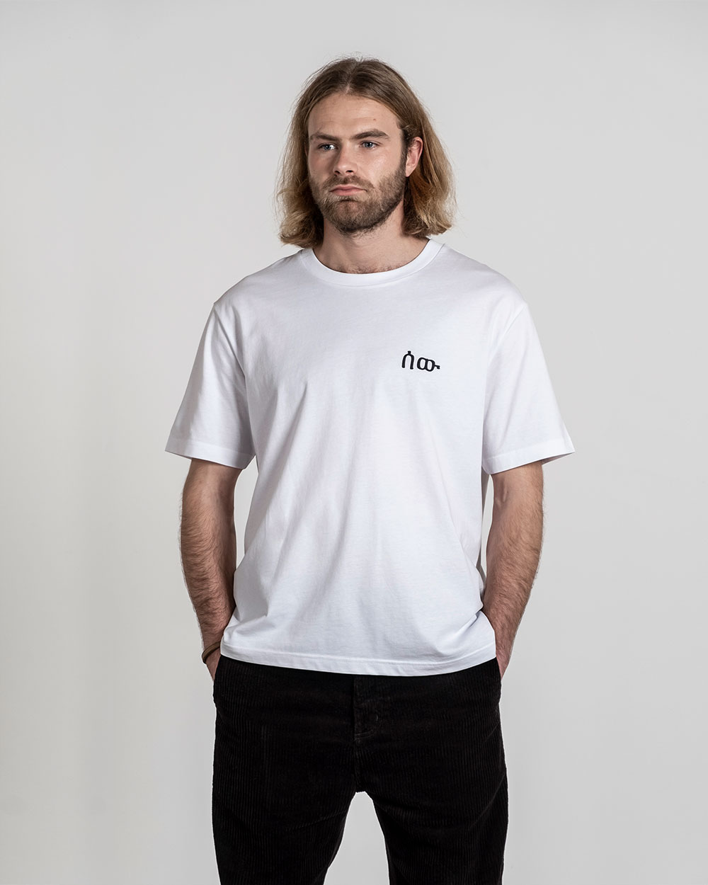 Weißes T-Shirt mit schwarzem Frontprint auf der linken Brust, getragen von einem stehenden Model mit den Händen in den Hosentaschen.