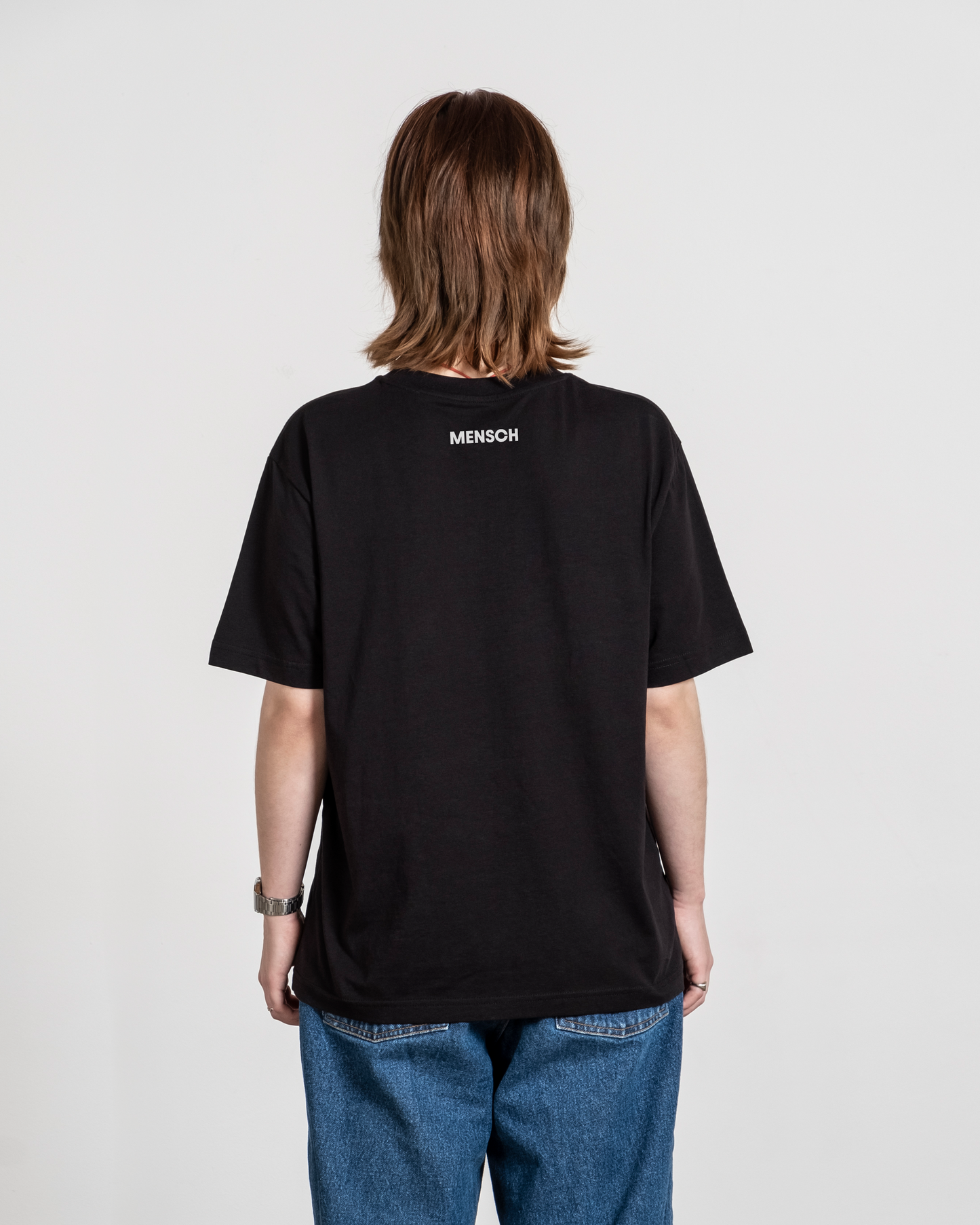 Schwarzes T-Shirt mit weißem „MENSCH“-Print im Nackenbereich, von hinten an einer Person getragen.