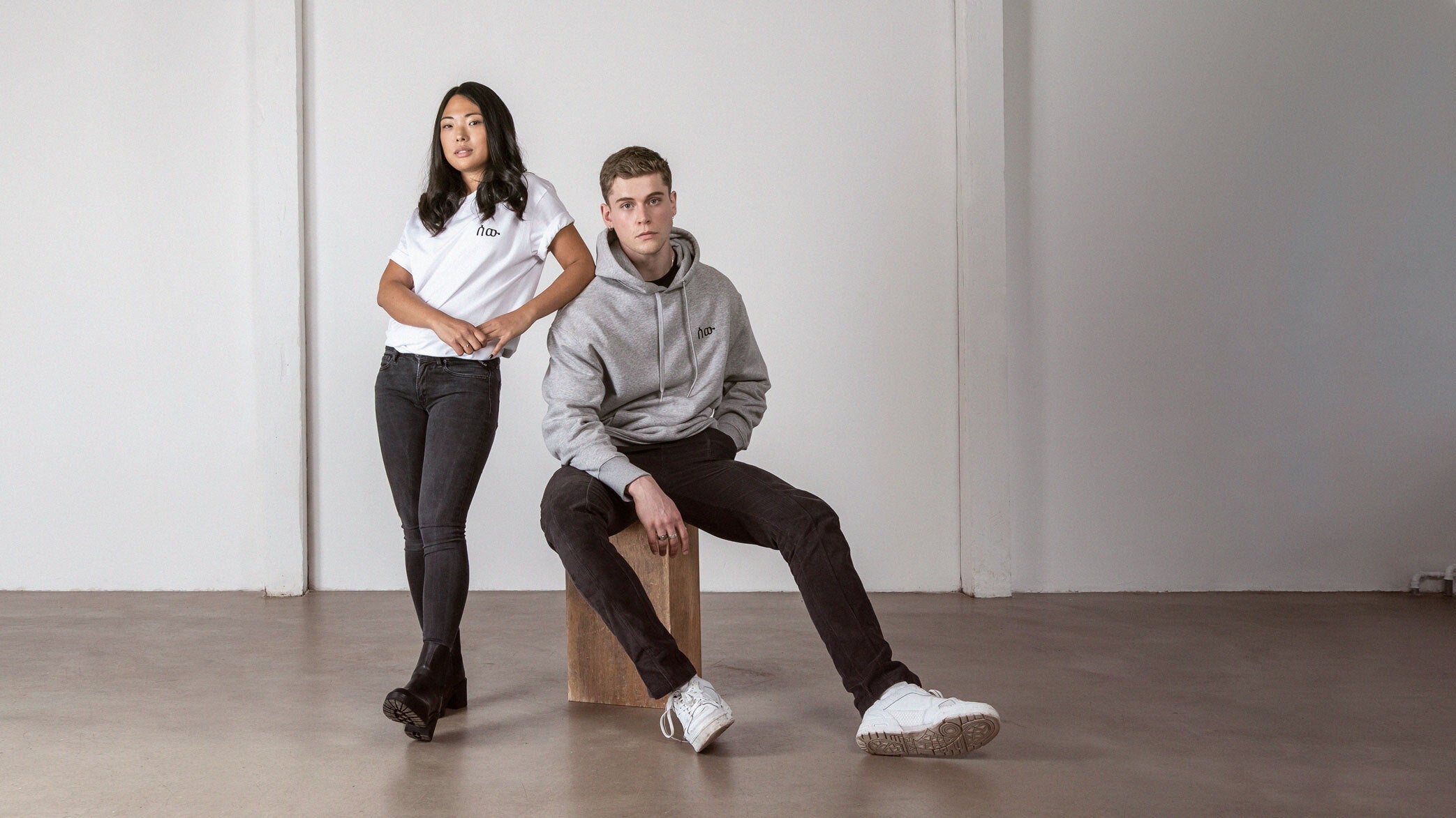 Ein junger Mann sitzt auf einem schlichten Holzhocker in einem hellen, leeren Raum mit Betonboden und weißer Wand. Er trägt einen grauen Hoodie mit schwarzem Logo und schwarze Jeans. Neben ihm steht eine junge Frau in weißem T-Shirt mit demselben Logo, dunkle