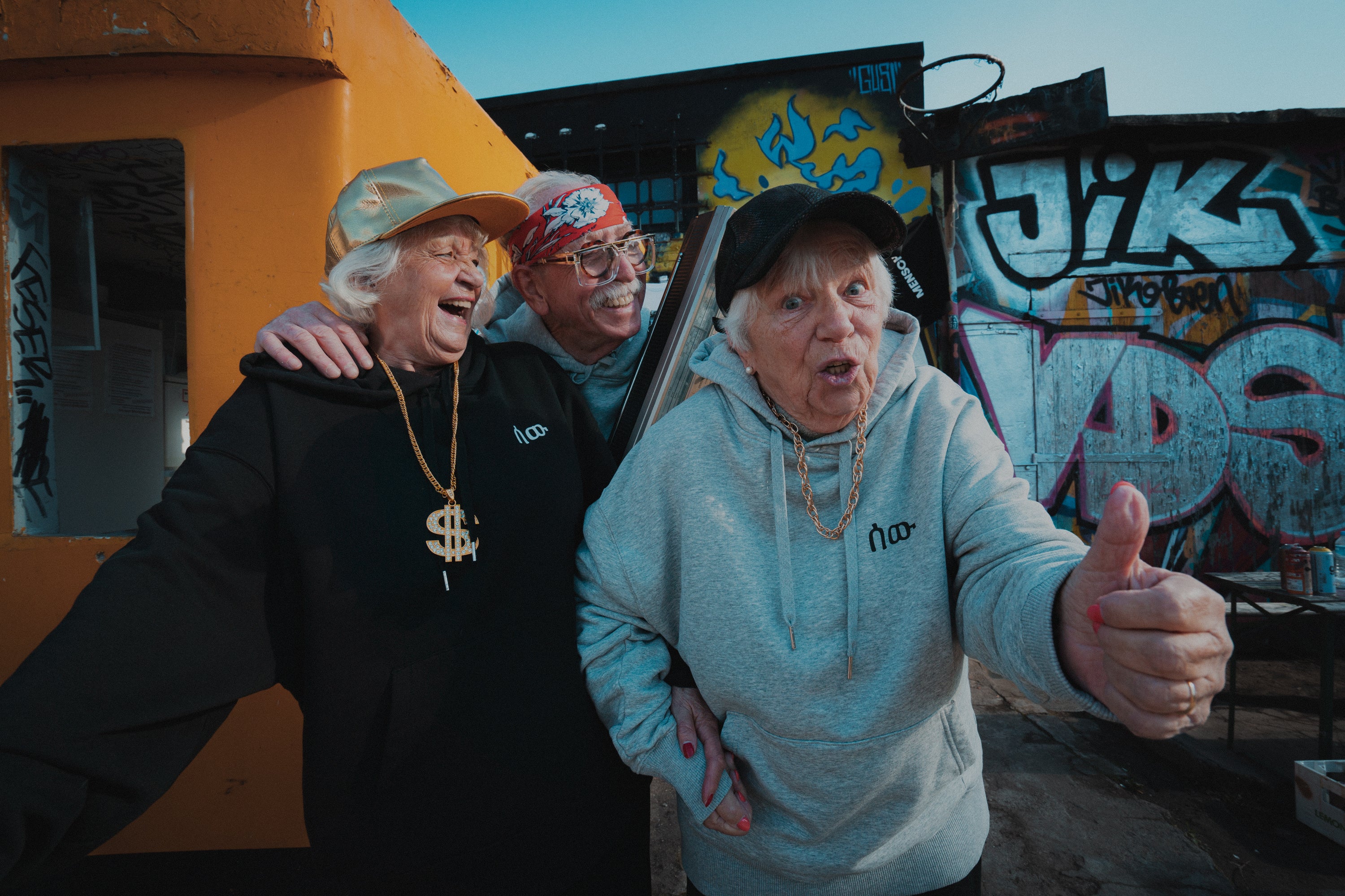 Drei ältere Menschen stehen fröhlich posierend vor einer bunt besprühten Graffiti-Wand. Zwei Frauen und ein Mann tragen lässige Streetwear-Hoodies. Die Frau links trägt einen schwarzen Hoodie, eine goldene Kappe und eine auffällige Dollarzeichen-Kette. Der Mann in der Mitte trägt eine rote Bandana und eine Brille. Die Frau rechts trägt einen grauen Hoodie und eine schwarze Cap, zeigt mit Daumen nach oben und hat eine goldene Kette um den Hals. Die Szene vermittelt Lebensfreude und Humor i