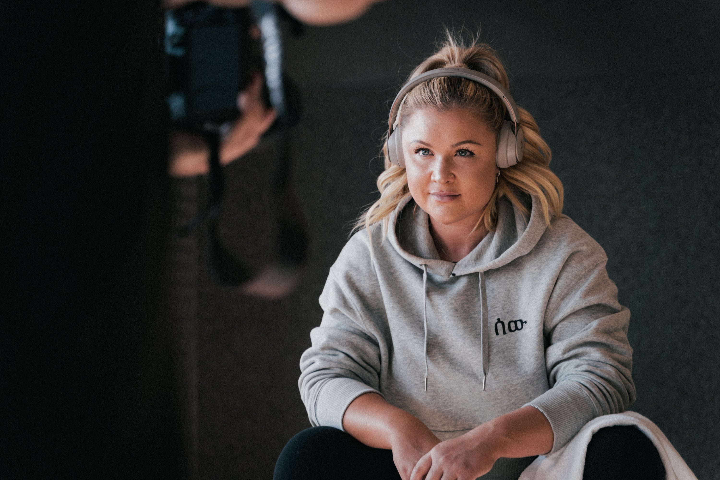 Eine junge Frau mit blonden, lockeren Haaren trägt große, ohrumschließende Kopfhörer und einen grauen Hoodie mit dem schwarzen Logo „∞oo“ auf der Brust. Sie sitzt entspannt mit verschränkten Armen und leicht geneigtem Kopf auf einem Hocker oder einer Bank. Im Vordergrund ist unscharf ein Kameramann zu sehen, der sie fotografiert oder filmt. Die Szene wirkt wie ein Blick hinter die Kulissen eines Shootings, aufgenommen in einem dezent ausgeleuchteten Innenraum.