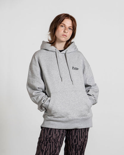 Grauer Kapuzenpullover mit lockerer Passform, Kängurutasche und schwarzem Frontprint eines minimalistischen Symbols auf der Brust, getragen von einer Person mit rötlich-braunen Haaren und dunkler gemusterter Hose.