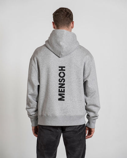 Grauer Hoodie mit großem schwarzem „MENSCH“-Print auf der Rückseite, getragen von einer Person mit kurz geschnittenem Haar; die Kapuze liegt auf dem Rücken.