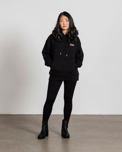 Vorderansicht eines schwarzen Hoodies mit weißer, minimalistisch gestickter Symbolschrift auf der linken Brust. Getragen von einer stehenden Person mit den Händen in der Kängurutasche.