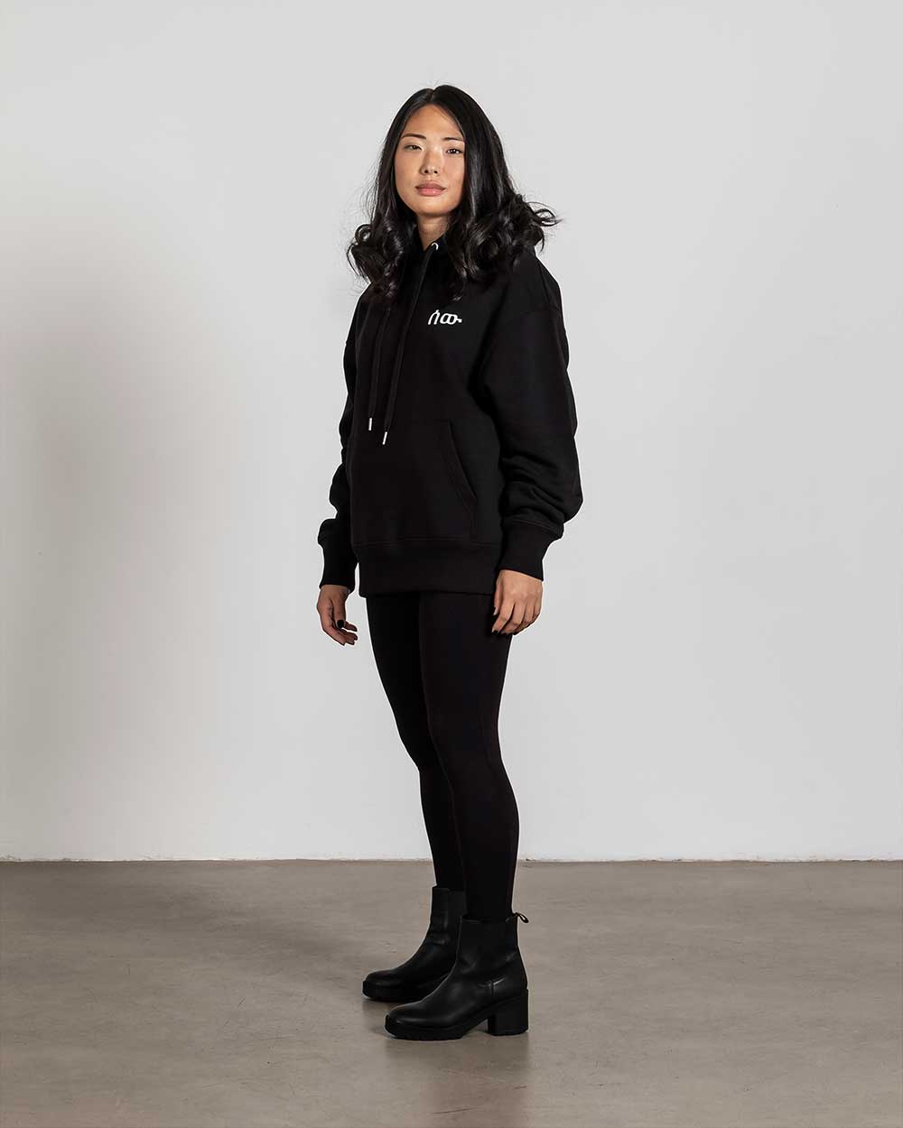Seitenansicht des schwarzen Hoodies mit Blick auf die weiße Bruststickerei und die leicht lockere Passform. Die Ärmel fallen über die Handgelenke.