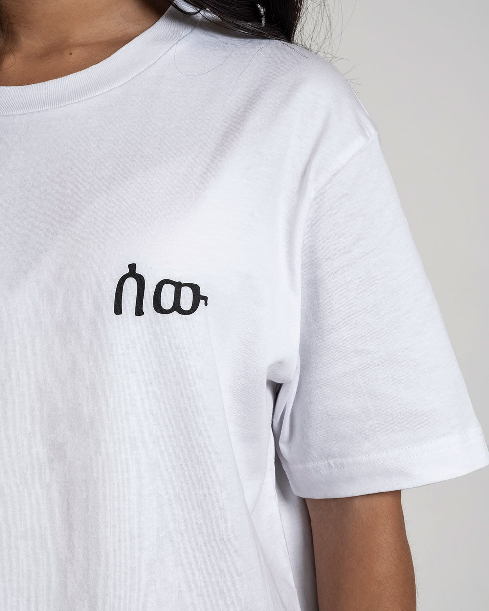Nahaufnahme des weißen T-Shirts mit schwarzem Frontprint auf der linken Brust – ein stilisierter, typografischer Schriftzug.