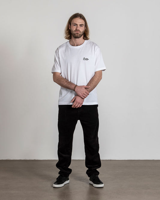 Weißes T-Shirt mit schwarzem Frontprint auf der linken Brust, getragen von einer Person mit langen Haaren und Bart, stehend mit verschränkten Armen.