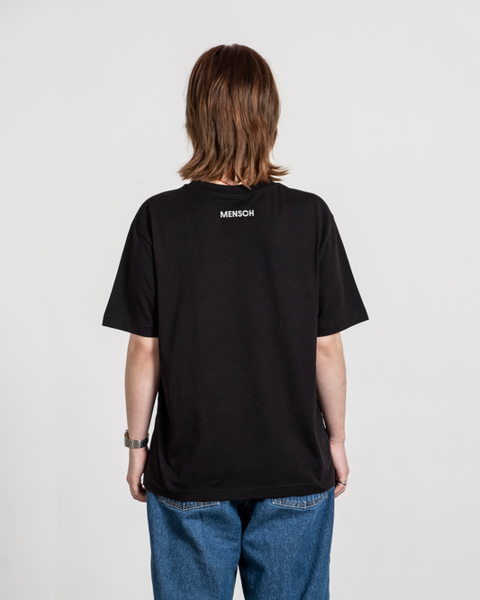 Schwarzes T-Shirt mit weißem „MENSCH“-Print im Nackenbereich, von hinten an einer Person getragen.